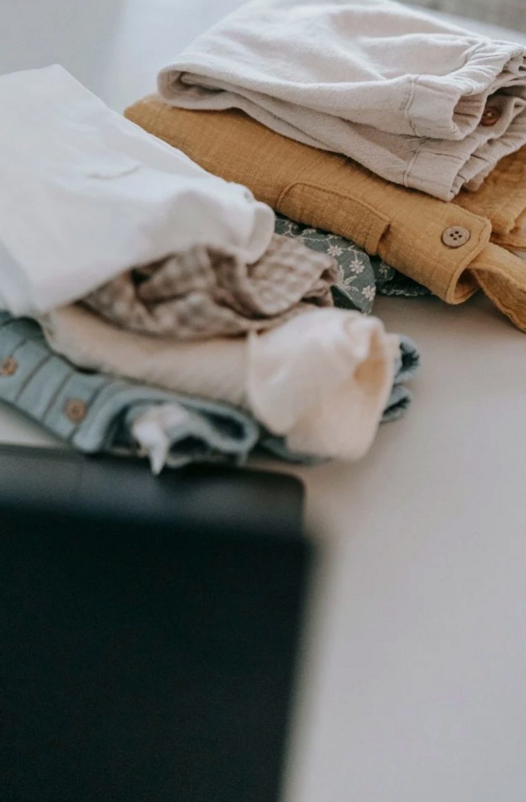 Abundance of colorful baby clothes folded on white clean table with opened modern blurred laptop in light room at home